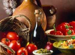 A basket of tomatoes and a bottle of olive oil beside bowls of peppers, olives, and salad ingredients.