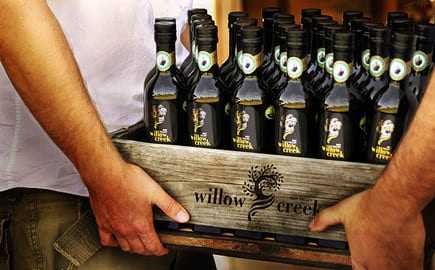 Hands carry a wooden crate of Willow Creek olive oil bottles.