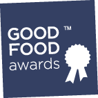 Logo of the Good Food Awards featuring a ribbon and text on a blue background. - Olive Oil Times