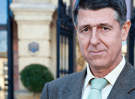 Man wearing a brown suit and a green tie, standing outdoors near a building entrance. - Olive Oil Times