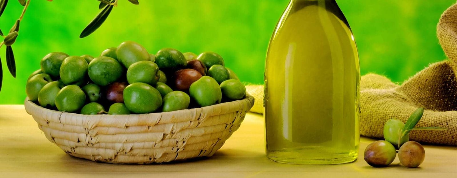A basket filled with green and brown olives next to a bottle of olive oil on a table. - Olive Oil Times