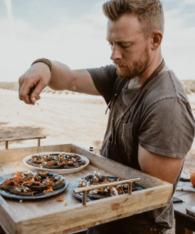 A man in a gray shirt garnishing plates of food on a wooden tray outdoors. - Olive Oil Times