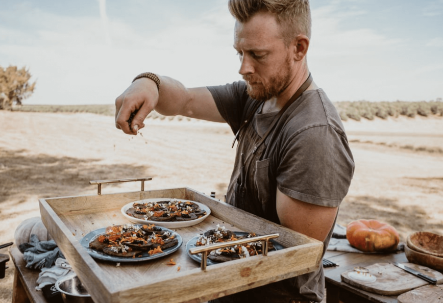 A man in a gray shirt garnishing plates of food on a wooden tray outdoors. - Olive Oil Times