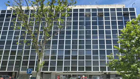 A modern office building featuring a grid pattern of large windows and a tree in front. - Olive Oil Times
