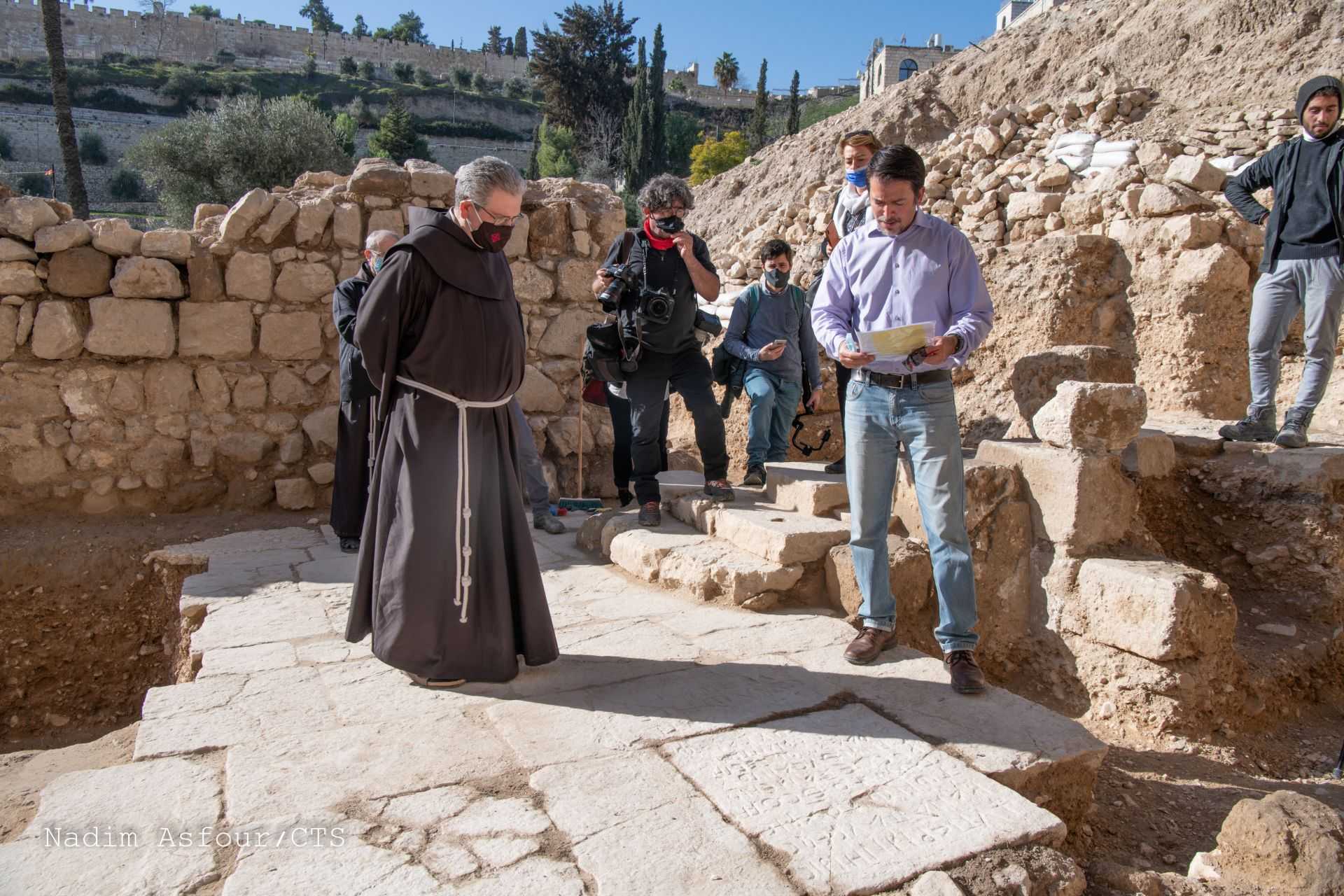 A monk observes an archaeologist reading from a document at a dig site, surrounded by several onlookers.