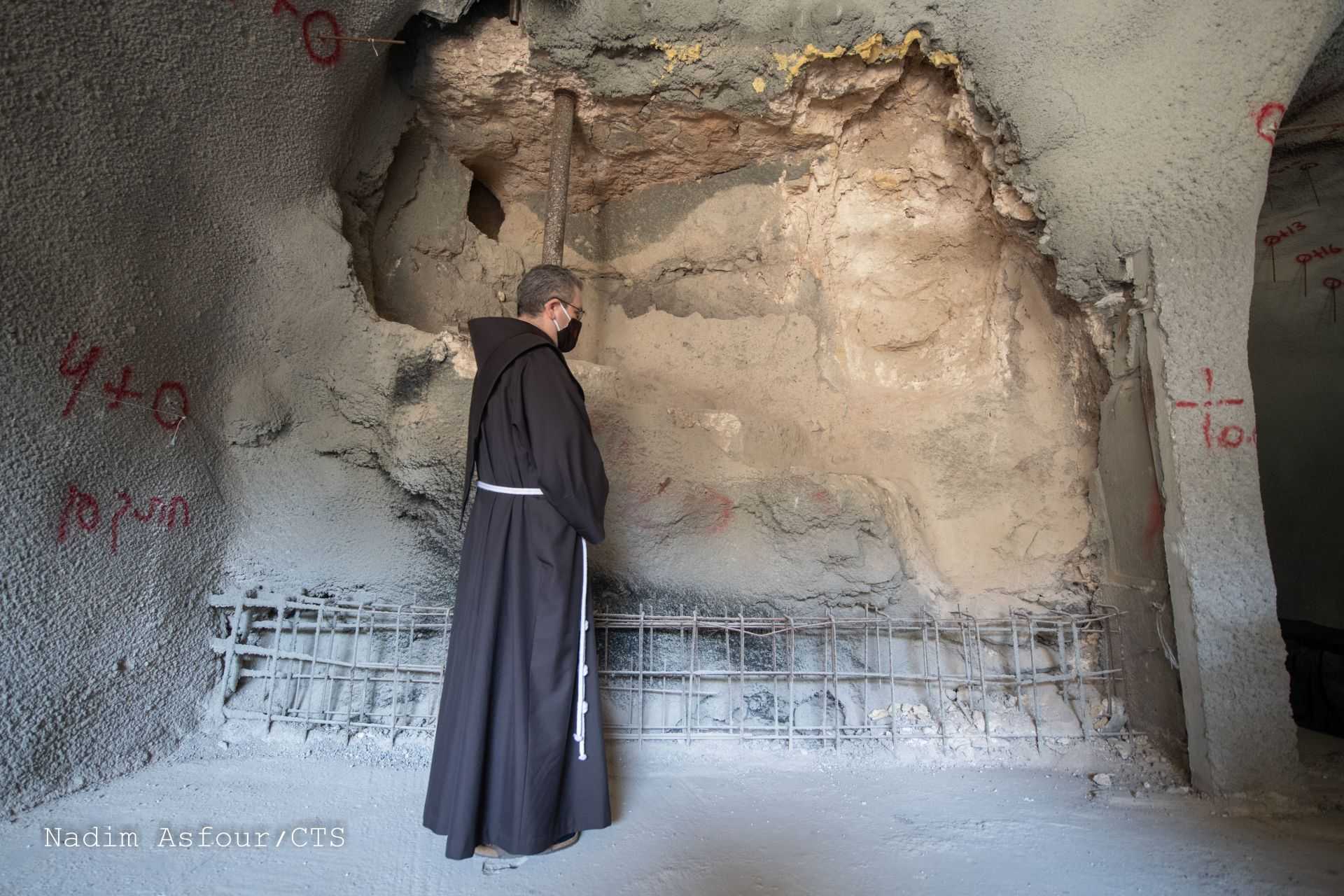 A monk inspects a damaged wall in a historical building, with markings visible on the surrounding area.