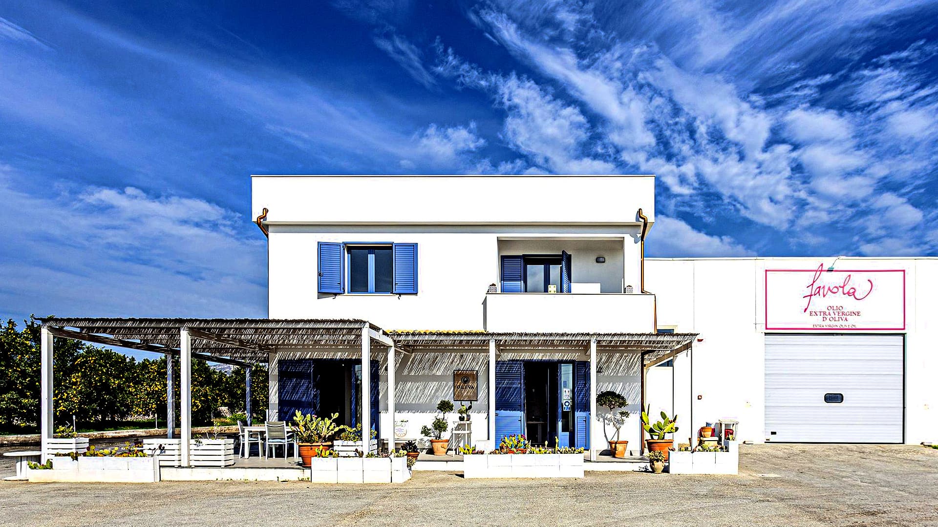 A white two-story building with blue shutters and a sign, surrounded by potted plants under a clear sky.