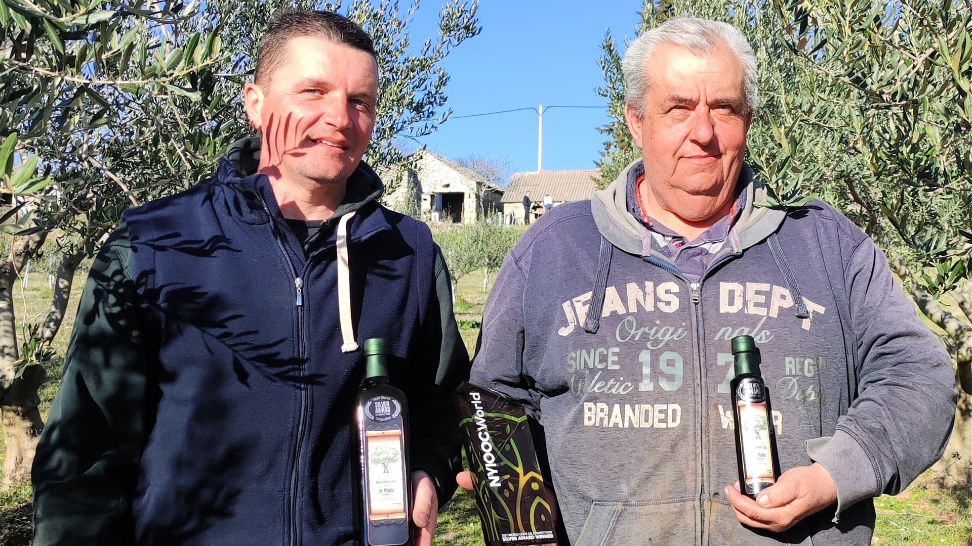 Two men in an olive grove hold bottles of olive oil, smiling at the camera.