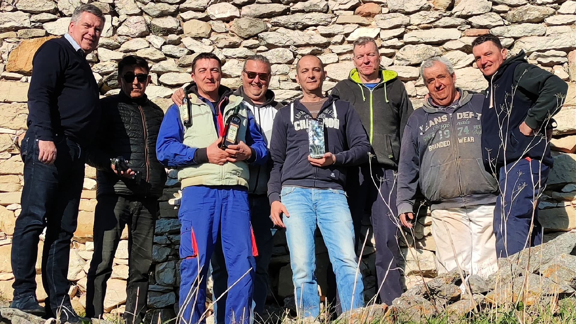 A group of eight men standing together, holding awards, in front of a stone wall.