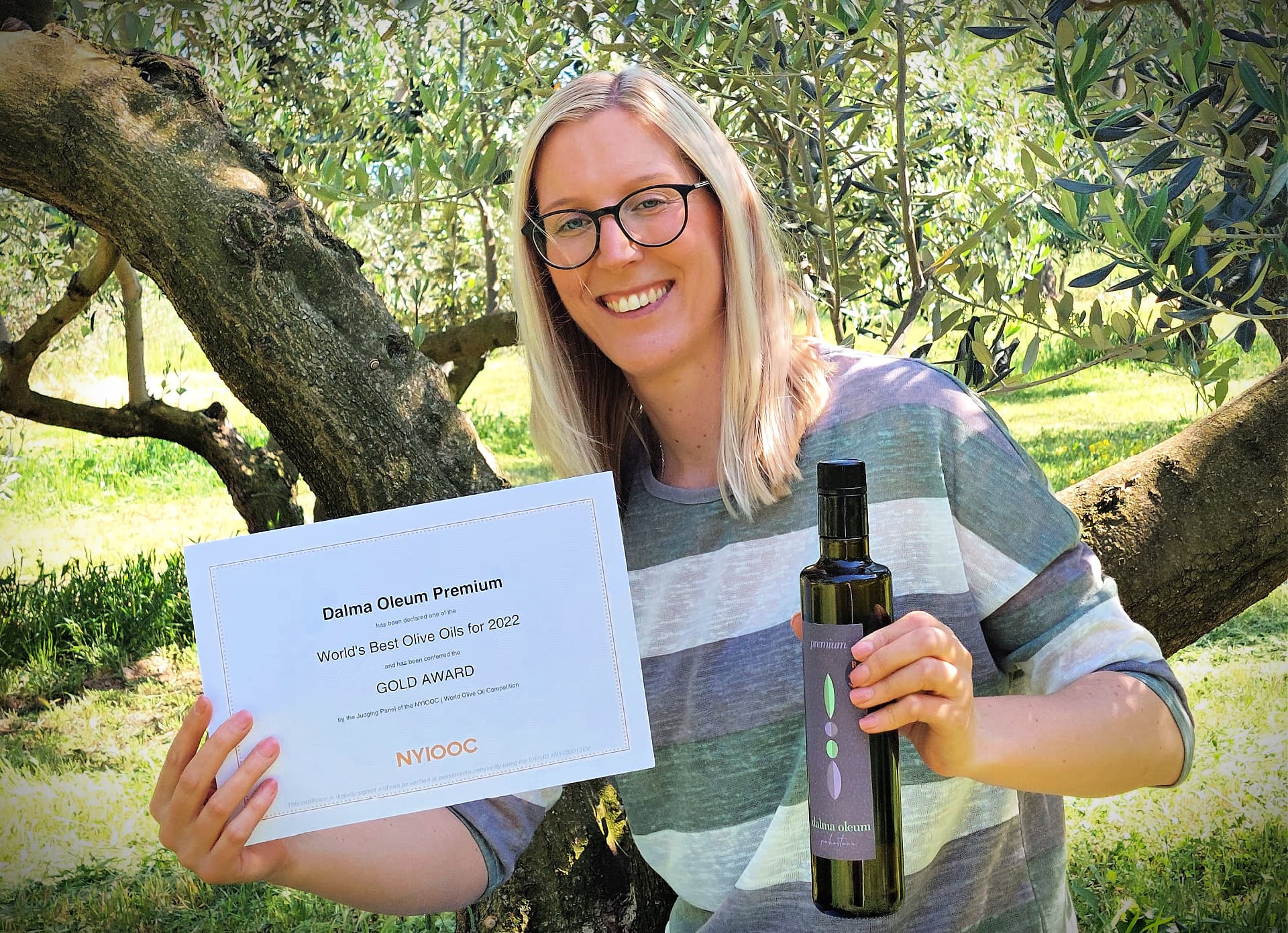 A woman holds a gold award certificate and a bottle of olive oil in an olive grove.