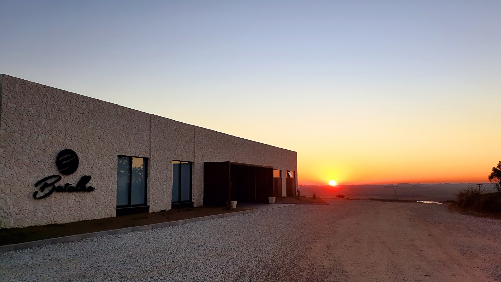 A modern building sits beside a gravel road at sunset, with orange hues in the sky.