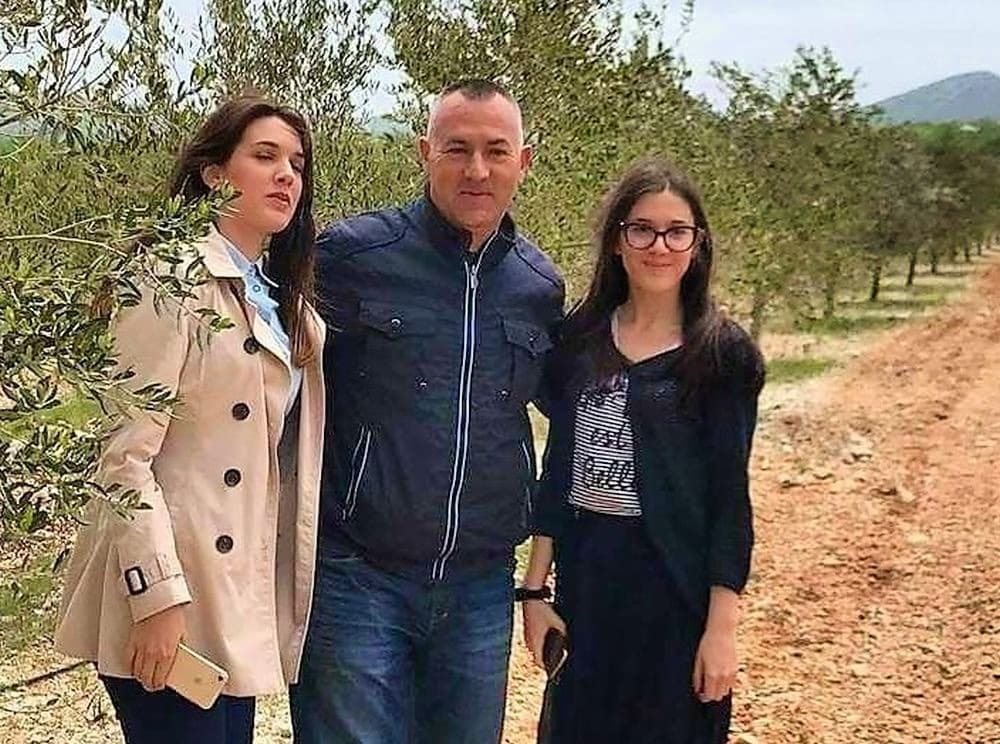 Three people pose for a photo among olive trees in a rural setting.