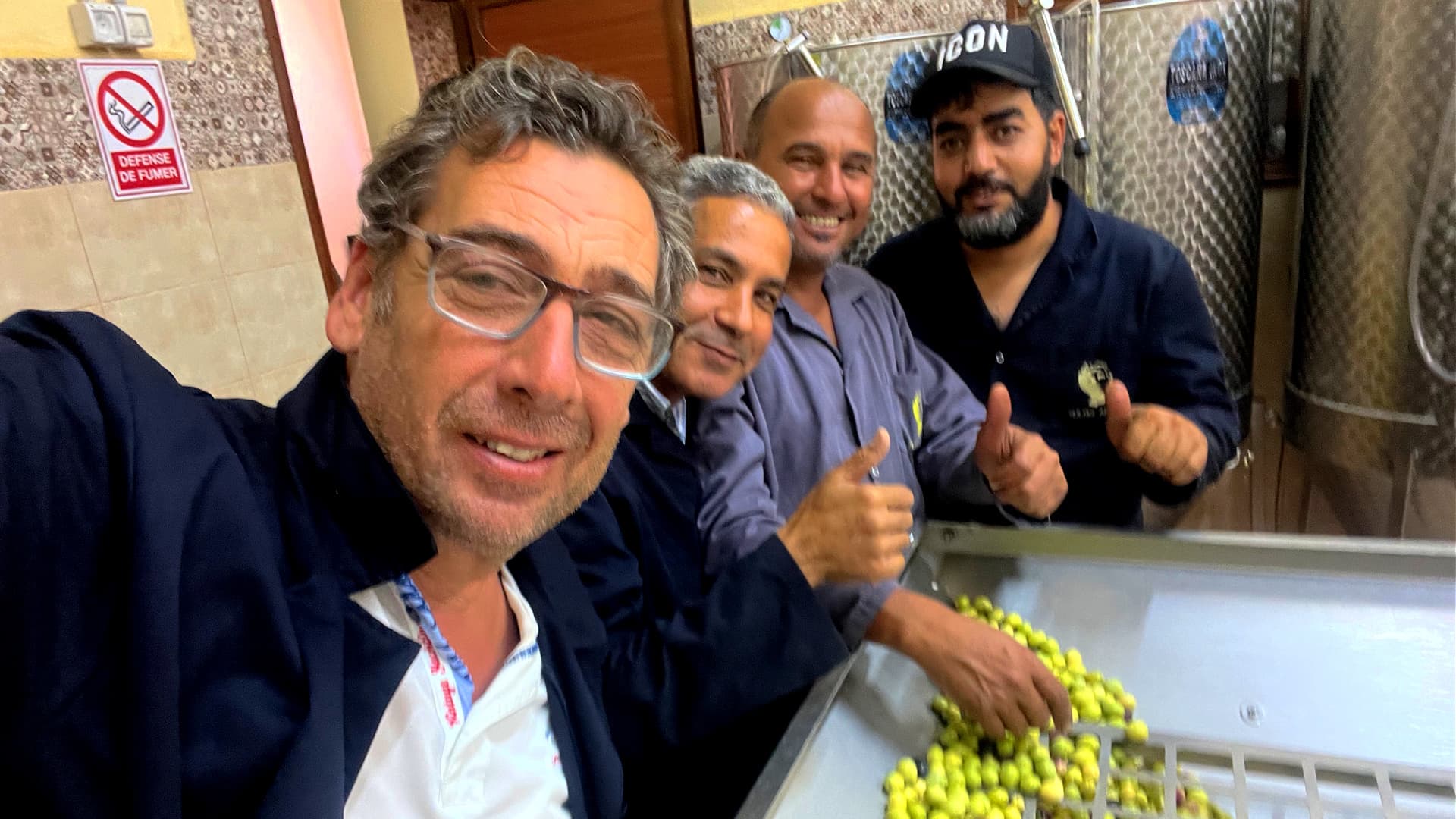 Four men smile and give thumbs up while working with green olives in a processing facility.