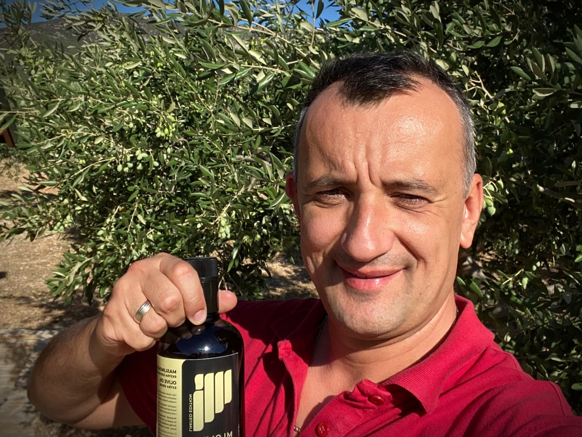 A man in a red shirt smiles while holding a bottle of olive oil in an olive grove.
