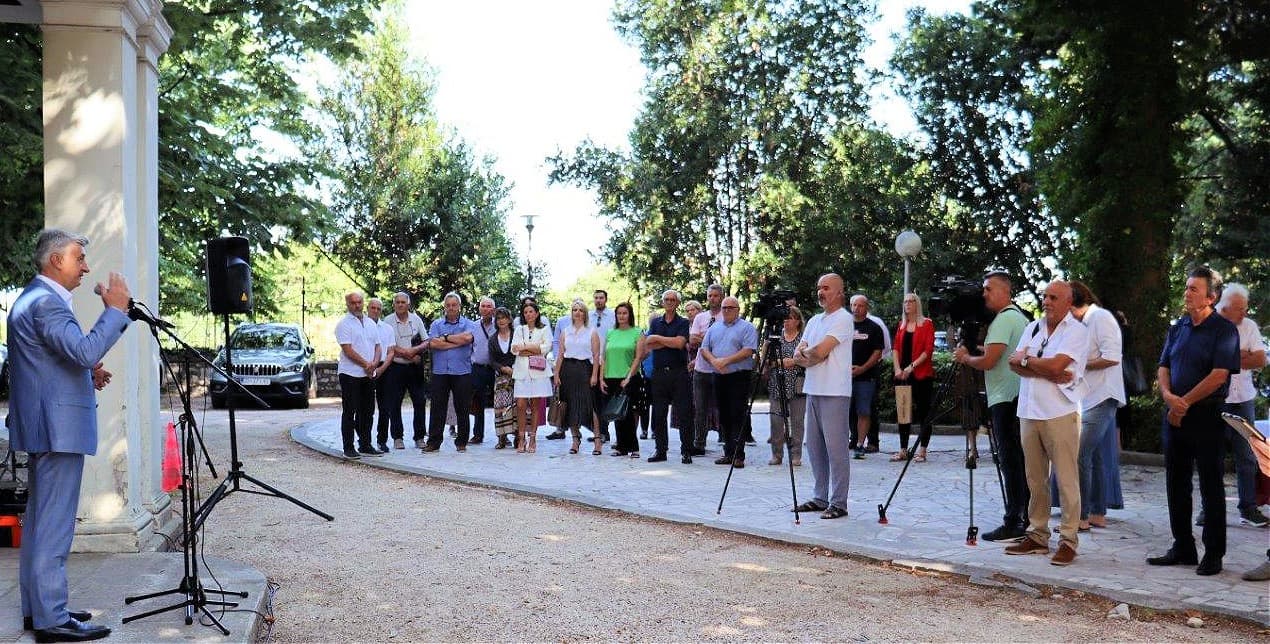 A speaker addresses a large crowd gathered outdoors among trees for a news event.