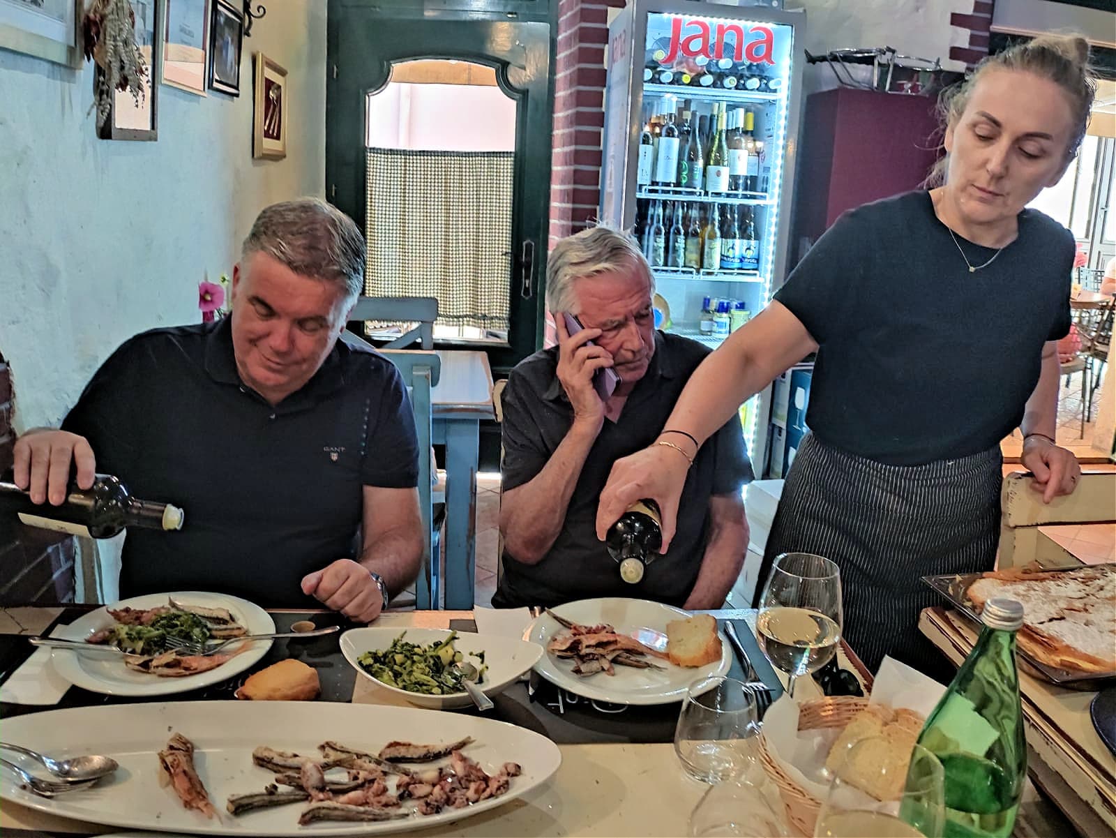 Two men dine at a table while a waitress pours wine for them in a cozy restaurant setting.