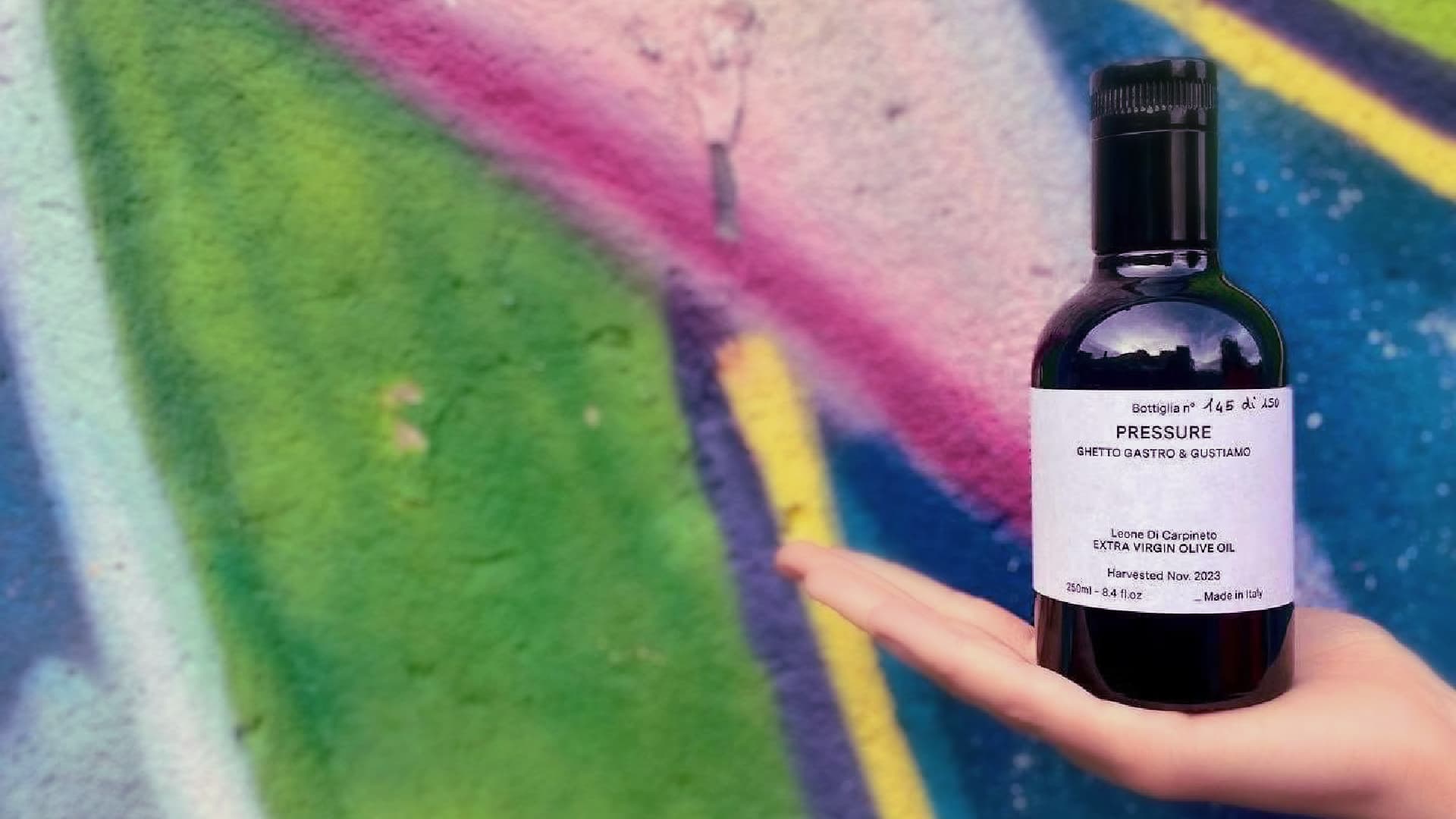 A hand holding a bottle of olive oil labeled "PRESSURE" against a colorful graffiti background.