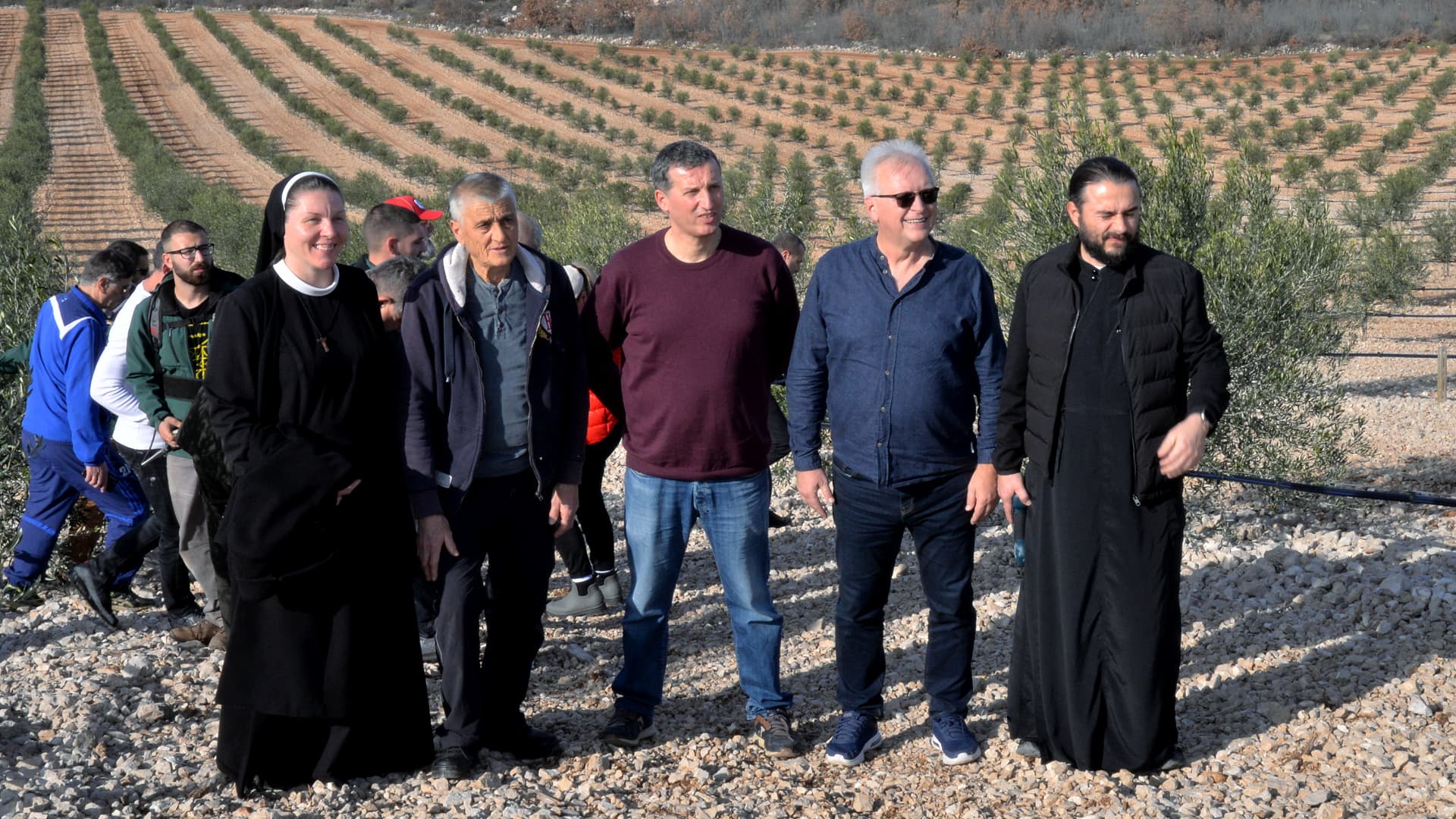 A diverse group of five people stands together in an olive grove with rows of trees in the background.