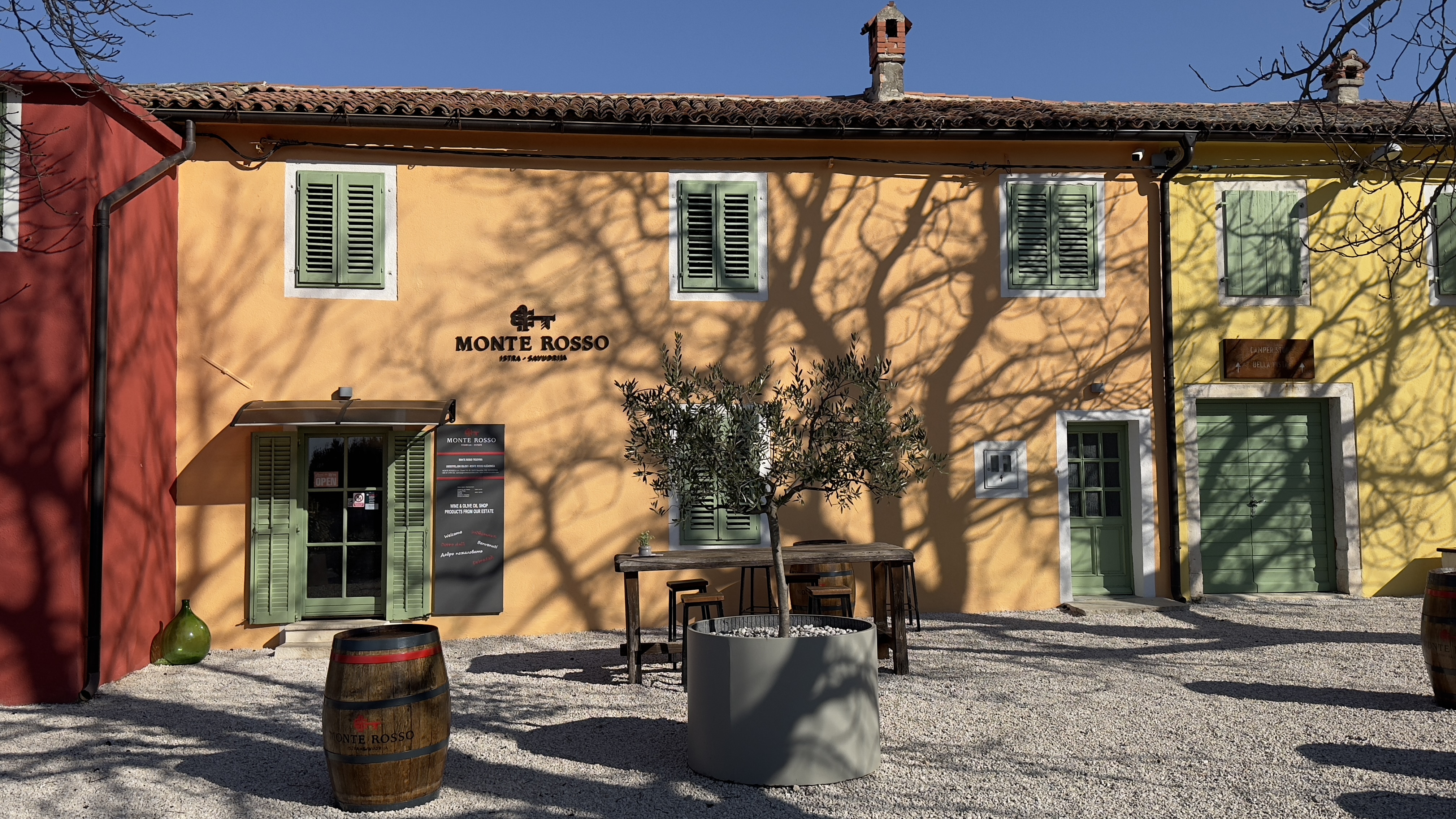Brightly colored buildings with green shutters and a wooden table in front, surrounded by olive trees and barrels.