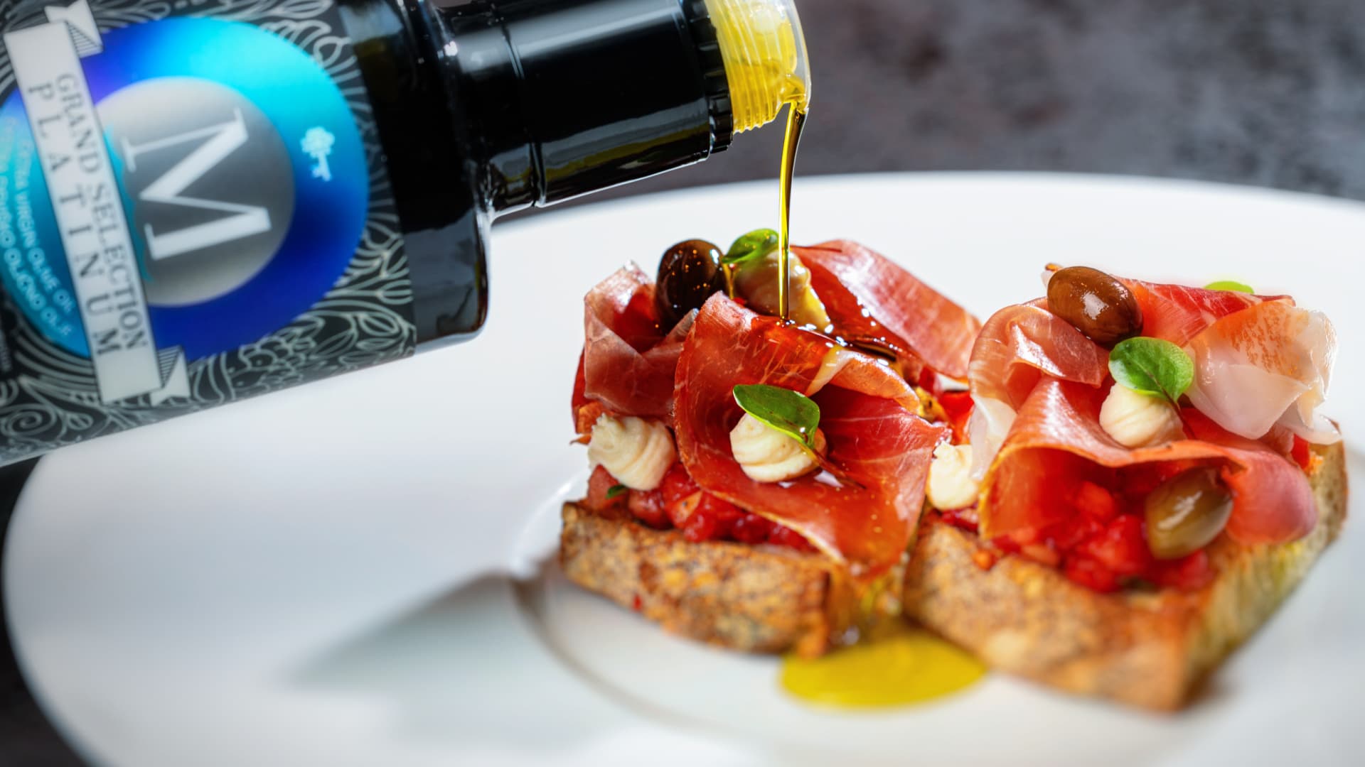 A bottle of olive oil drizzles over bruschetta topped with prosciutto, olives, and herbs.