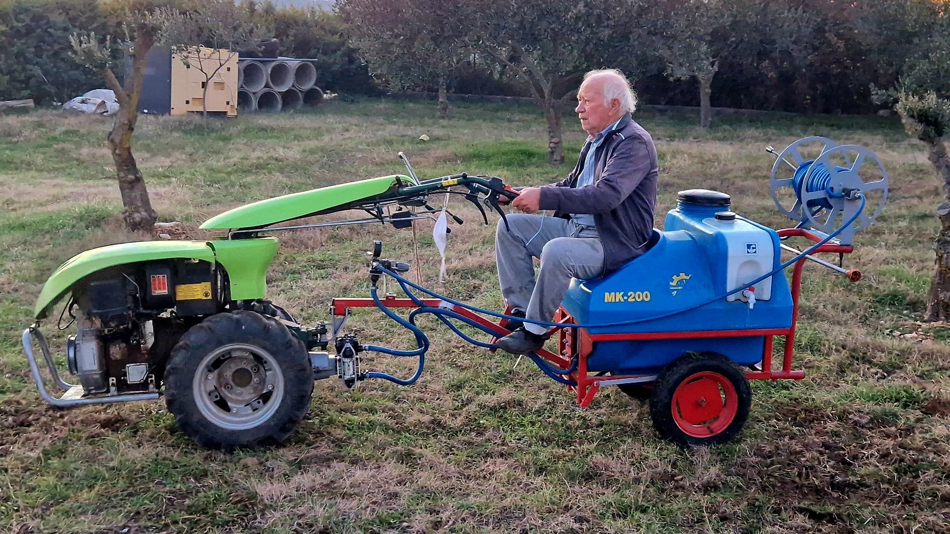 An elderly man operates a small tractor pulling a sprayer in an orchard.